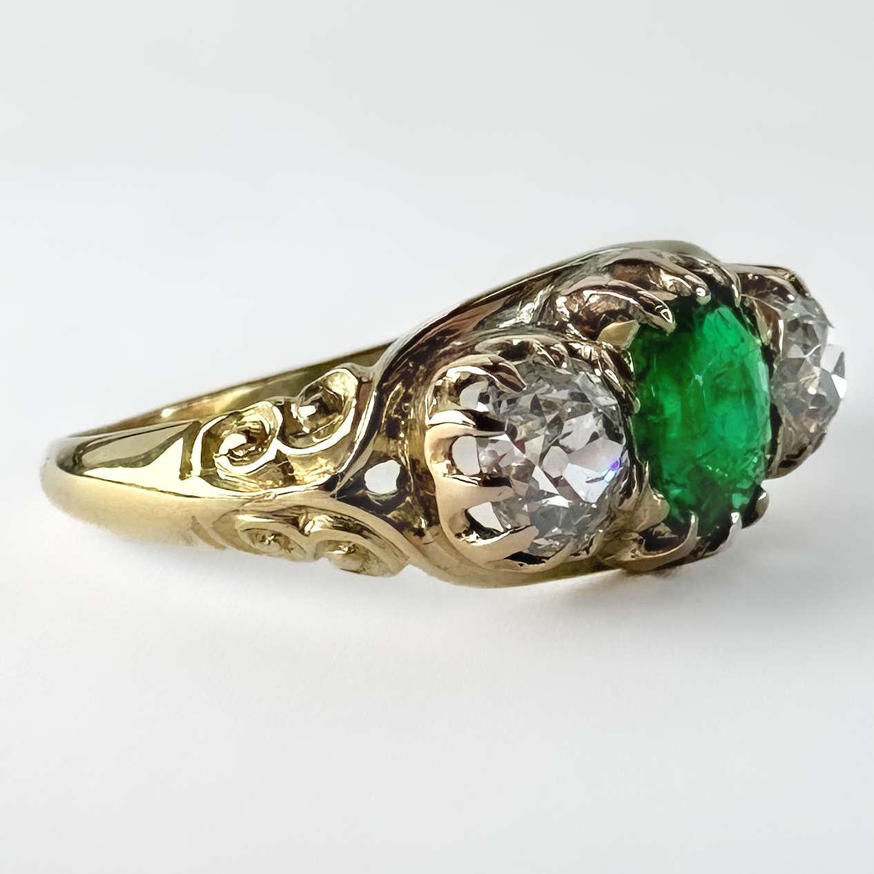 Antique Emerald Diamond 3 Stone Ring. This beautiful victorian ring has 3 claw set stones in 18ct yellow gold. The center emerald stone in a vivid green shade and weighs 1ct. This nice bright stone is faceted cushion cut. Either side of the emerald is two cushion shaped victorian brilliant cut diamonds weighing 0.6cts each. The 18ct yellow gold ring features a carved gallery and carved scrolled shoulders. The ring sits low on the finger. Please contact us if you would like to know more about this ring or visit us at Saddingtons Jewellery, Mayfair.