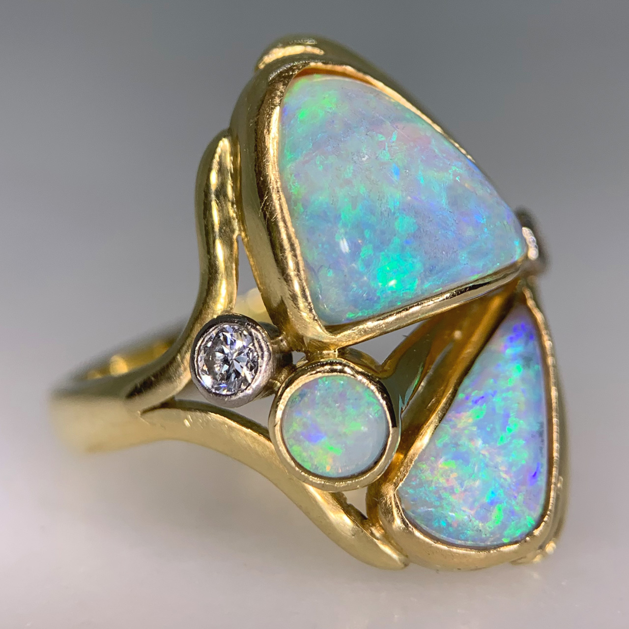 Fancy Asymmetric Water Opal and Diamond Ring in 18ct Yellow Gold, Hallmarked London, 1963. The unusual vintage custom made ring holds three vibrant water opals, two are of asymmetric shape, one circular. The opals are a mesmerising turquoise blue and purple with hints of green and yellow, we would even go as far as saying the colours are fluorescent. The split shoulders are decorated with two brilliant cut diamonds, of different cartage. The total head of the ring measures approximately 21.8 mm x 17 mm, The depth of the head 7 mm approximately. All the stones are bezel set in a half round polished shank. This ring is in immaculate condition, the opal will be professionally polished before any purchase. 