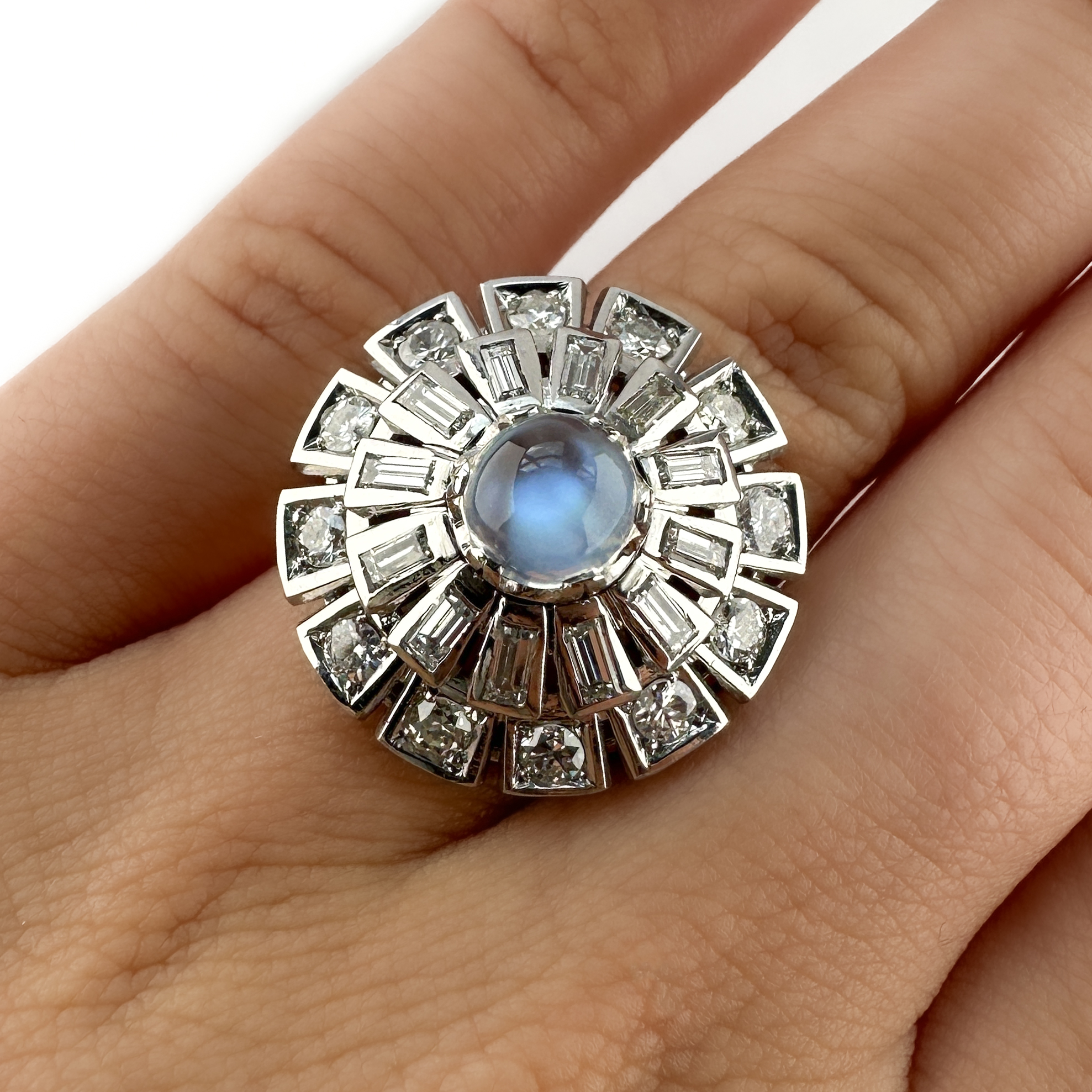 Splendid 1960's Diamond & Moonstone Cocktail Ring. This striking designer 1960s platinum cocktail ring is centred with a luminous oval moonstone, displaying a soft blue that shifts beautifully in the light. Surrounding the moonstone is a art-deco inspired architectural arrangement of 12 round brilliant-cut and 12 baguette cut diamonds, creating a dramatic sunburst effect synonymous with mid-century design. The strong geometric structure and sculptural dome form give the ring an unmistakably glamorous presence on the finger. The ring distinguished by its sculptural platinum mount, featuring a finely fluted shank that draws the eye towards the setting. Beneath the head, an intricate openwork gallery with scrolling pierced detail provides both lightness and architectural depth to the ring. On the outside of the platinum shank there is a makers mark. The bright diamonds are G in colour and VS1 clarity with a total weight of 1.8ct. This ring can be resized. Please contact us if you would like to know more about this stunning ring or visit us at Saddingtons Jewellery, Mayfair.