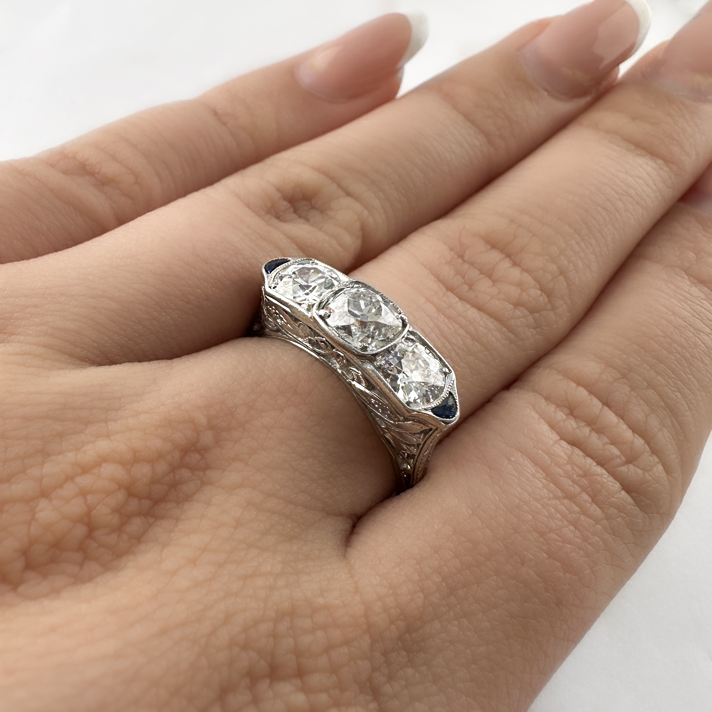 Fabulous Art Deco 2.5ct Diamond Trilogy Ring. This ring is an incredible example of art deco design with its beautiful ornate pierced gallery that features scrolls and florals. The top features 3 large sparkly old transitional cut natural diamonds. The centre diamond is 1.17cts with the total weight being 2.5ct. The diamond are H in colour and VS2 clarity. The diamonds are accented by two deep blue sapphires. Its elongated geometric design is very flattering on the finger. The stones are set in platinum and have milgrain edges. The carved shoulders taper elegantly into a slender round band. This ring can be resized. Please contact us if you would like to know more about this amazing ring or visit us at Saddingtons Jewellery, Mayfair.