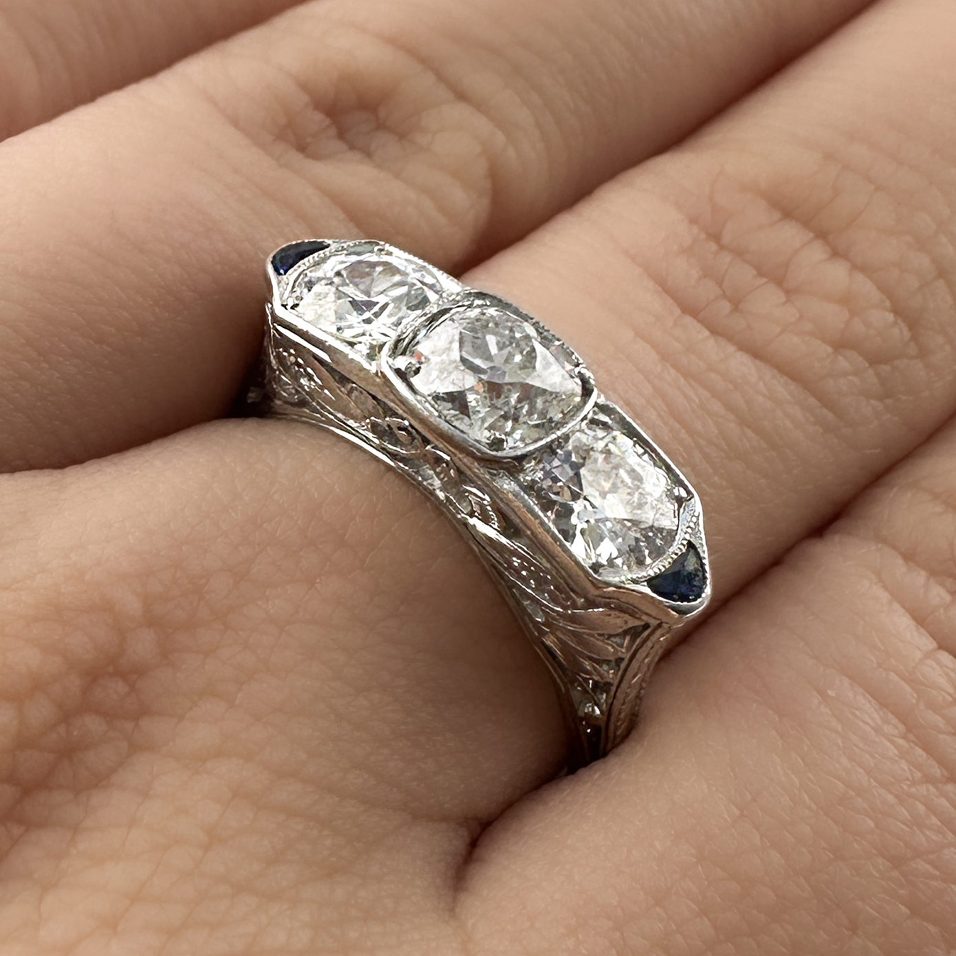 Fabulous Art Deco 2.5ct Diamond Trilogy Ring. This ring is an incredible example of art deco design with its beautiful ornate pierced gallery that features scrolls and florals. The top features 3 large sparkly old transitional cut natural diamonds. The centre diamond is 1.17cts with the total weight being 2.5ct. The diamond are H in colour and VS2 clarity. The diamonds are accented by two deep blue sapphires. Its elongated geometric design is very flattering on the finger. The stones are set in platinum and have milgrain edges. The carved shoulders taper elegantly into a slender round band. This ring can be resized. Please contact us if you would like to know more about this amazing ring or visit us at Saddingtons Jewellery, Mayfair.