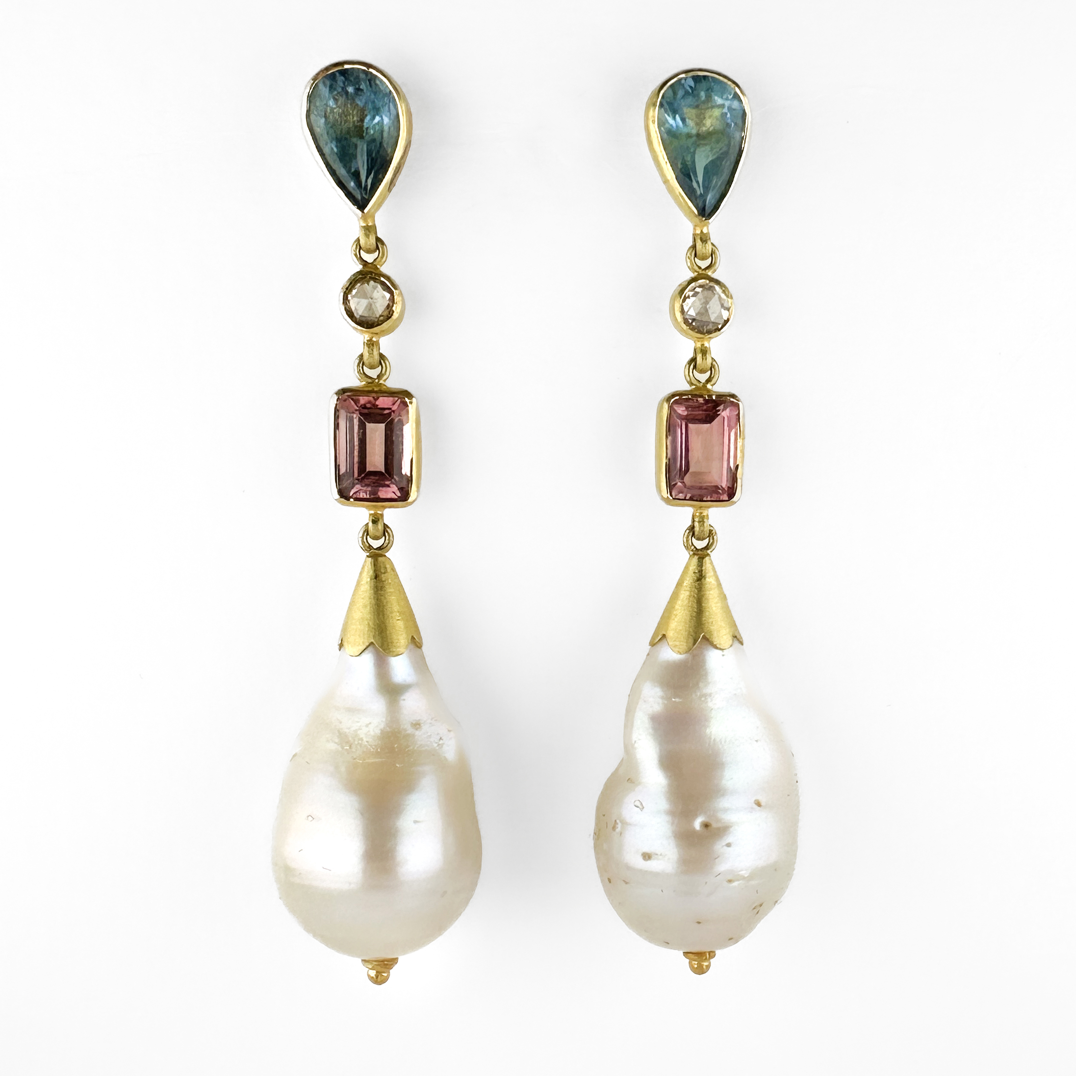 Impressive Multi-Gemstone & Baroque Pearl Drop Earrings. An exceptional pair of artisan-crafted gold drop earrings, combining luminous baroque natural pearls with an elegant arrangement of coloured gemstones in richly contrasting tones. The design begins with pear-shaped blue gemstones at the stud, suspended beneath by delicate articulated links accented with small round stones and emerald-cut pink tourmaline, culminating in substantial baroque pearl drops. All the stones are mounted in 18ct yellow gold with a matte finish. The irregular natural form of the pearls gives the earrings an organic, sculptural beauty, while the geometric gemstone settings create a refined architectural balance. The soft lustre of the pearls contrasts beautifully against the vivid pink and deep blue stones, producing a composition that feels both luxurious and highly individual. The pearl and its fitting measure 30mmx14mm with the total length being 59mm. Elegant movement, asymmetry of texture and sophisticated colour layering give these earrings a distinctly one-of-a-kind atelier aesthetic. Dramatic yet wearable, they have the feel of contemporary studio jewellery. Please contact us if you would like to know more about these stunning earrings or visit us at Saddingtons Jewellery, Mayfair.