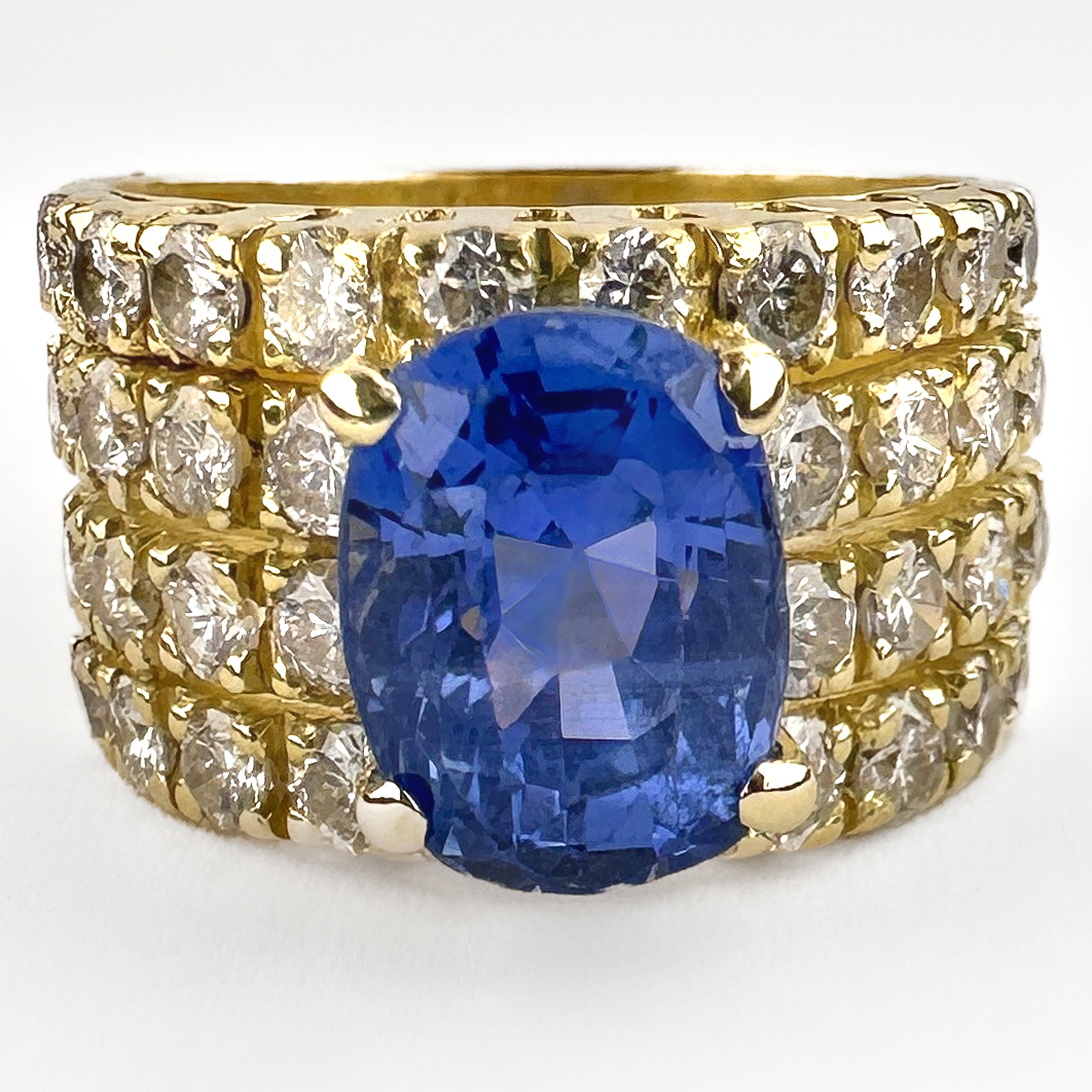 Stunning Ceylon Sapphire & Pavé Diamond Cocktail Ring. This amazing ring features a central natural unheated Ceylon sapphire that is claw set amongst a carpet of 36 pavé set natural diamonds. The total diamond weight is 0.9cts and they are round brilliant cut. The 2.5ct sapphire is a stunning cornflower, mid blue shade. The 18ct yellow gold 4 band design creates a chunky and interesting wide band that is very comfortable on. The gallery features lattice style open work. On the back of the ring there is a 750 stamp. Please contact us if you would like to know more about this beautiful ring or visit us at Saddingtons Jewellery, Mayfair.