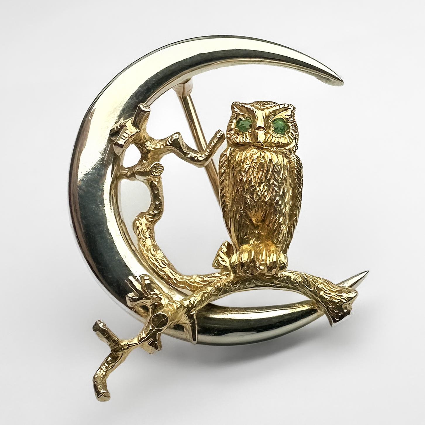 Vintage Gold Owl In The Moon Brooch. This sweet brooch depicts an owl on a branch in the crescent moon. It is made from 9ct white and yellow gold and has two emeralds for the eyes. On the back of the owl there is a 1987 Birmingham hallmark and a makers mark above. The brooch measures at 25mm in diameter and weighs 6.7 grams. Please contact us if you would like to know more about this beautiful brooch or visit us at Saddingtons Jewellery, Mayfair.