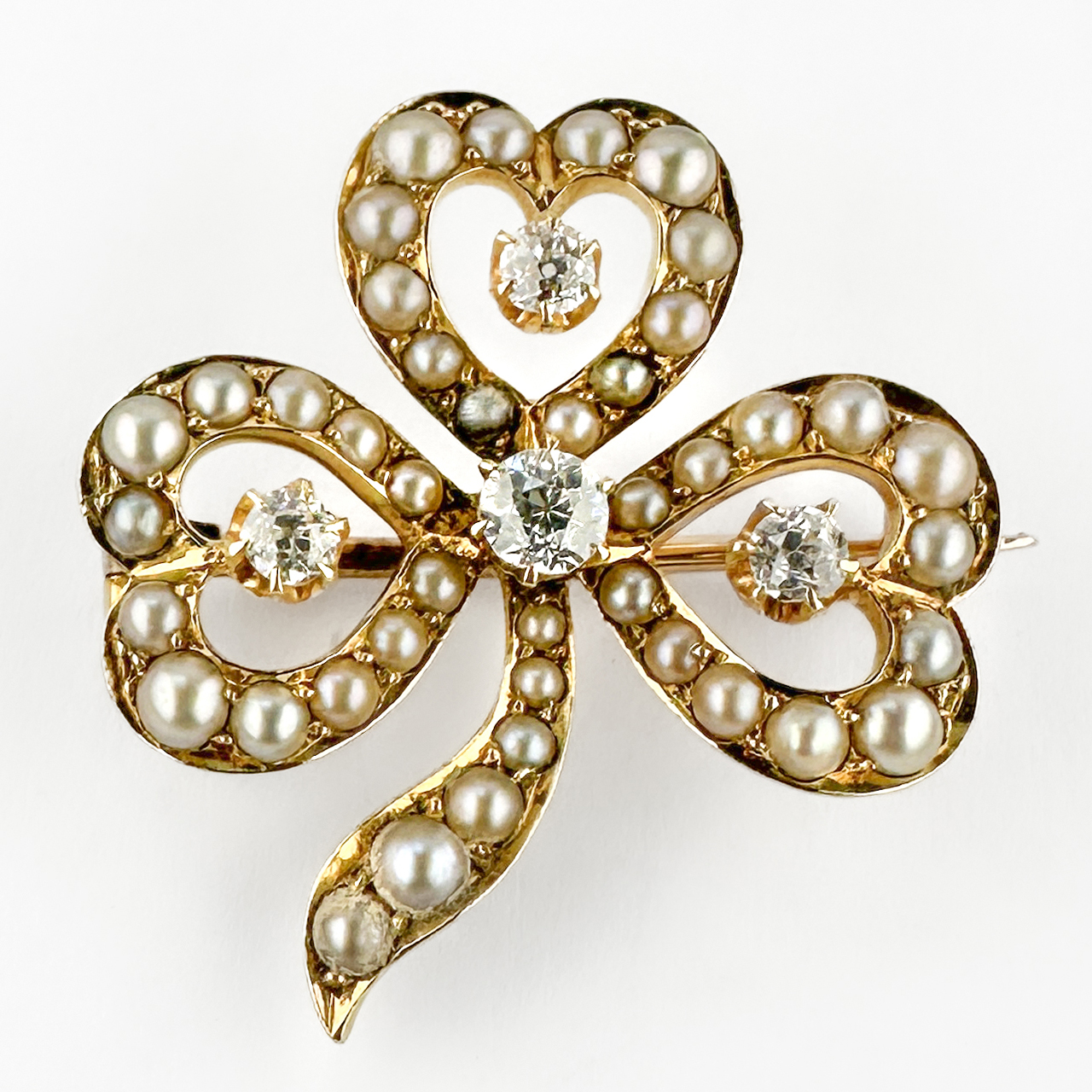 Edwardian Pearl & Diamond Clover Brooch. Stunning clover brooch with 42 pearls and 4 diamond set in 15ct yellow gold. The leaf heart shape sections have 12 pearls with a small transitional brilliant cut diamond claw set in the centre, the biggest diamond sits in the middle of the clover design. The openwork design creates a delicate lightness typical of refined late Victorian and Edwardian jewellery. Rich in symbolism, clover jewellery traditionally represented luck, hope, faith and love, making pieces such as this highly cherished sentimental gifts during the Victorian era. The soft glow of the pearls contrasts beautifully with the lively brilliance of the diamonds, creating an elegant and timeless composition.From top to bottom the brooch measures 2.8cm and it is 2.6cm wide. Please contact us if you would like to know more about this wonderful brooch or visit us at Saddingtons Jewellery, Mayfair.