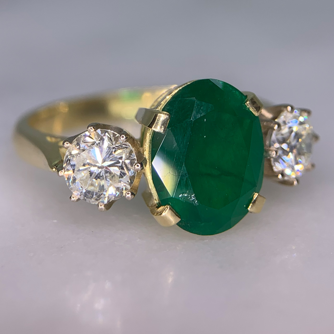 Exquisite Emerald and Diamond Trilogy Ring, in 18ct hallmarked yellow Gold, Birmingham 1996.  The centre deep emerald measures  11 mm x 9 mm and is enclosed by two diamonds totalling at 1.10carats of diamond weight. The total ring head measures 20 mm in width. All the stones are claw set with a basket depth of 6mm. The shank is polished with tiffany shoulders.  Excellent ring.