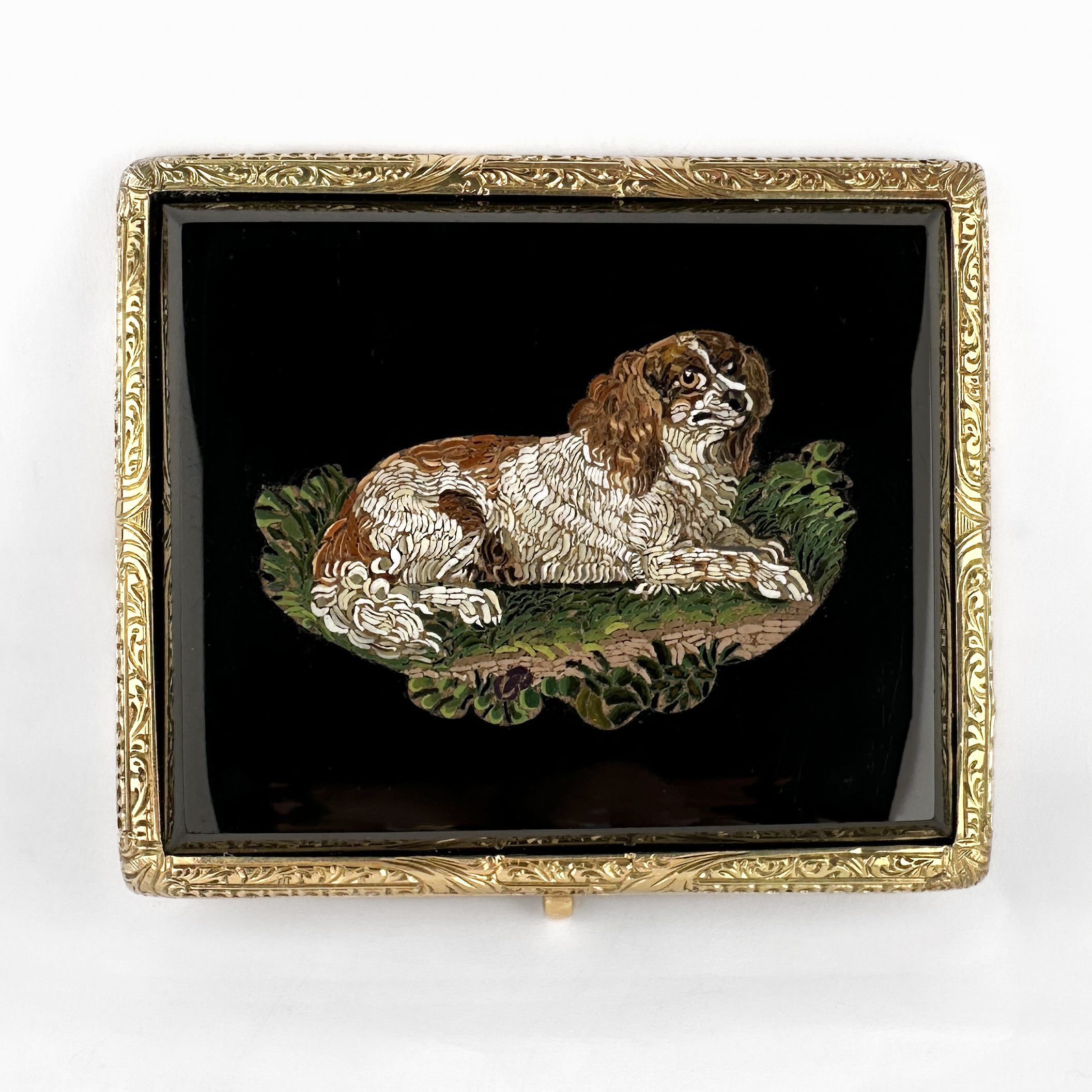 Victorian Dog Mosaic Brooch. An exceptional antique Victorian micro mosaic brooch crafted in 18ct gold, dating from the mid to late 19th century. The rectangular plaque is finely decorated with an exquisitely detailed micro mosaic depicting a reclining King Charles spaniel-type dog within a naturalistic landscape. Executed using incredibly small hand-set glass tesserae, the mosaic displays remarkable precision and artistry, particularly in the intricate rendering of the dog’s fur and surrounding foliage. Set against a rich black ground, the colourful mosaic composition is framed within an elegant nicely engraved 18ct gold mount that enhances the refinement of the piece without detracting from the extraordinary craftsmanship. Micro mosaic jewellery of this quality was highly sought after during the Victorian Grand Tour period, when affluent travellers acquired finely worked Roman mosaics as sophisticated souvenirs and collectors’ pieces. Animal subjects, particularly dogs, were especially admired for their symbolism of loyalty and companionship. The brooch measures 4x4.7cm. Please contact us if you would like to know more about this wonderful brooch or visit us at Saddingtons Jewellery, Mayfair.