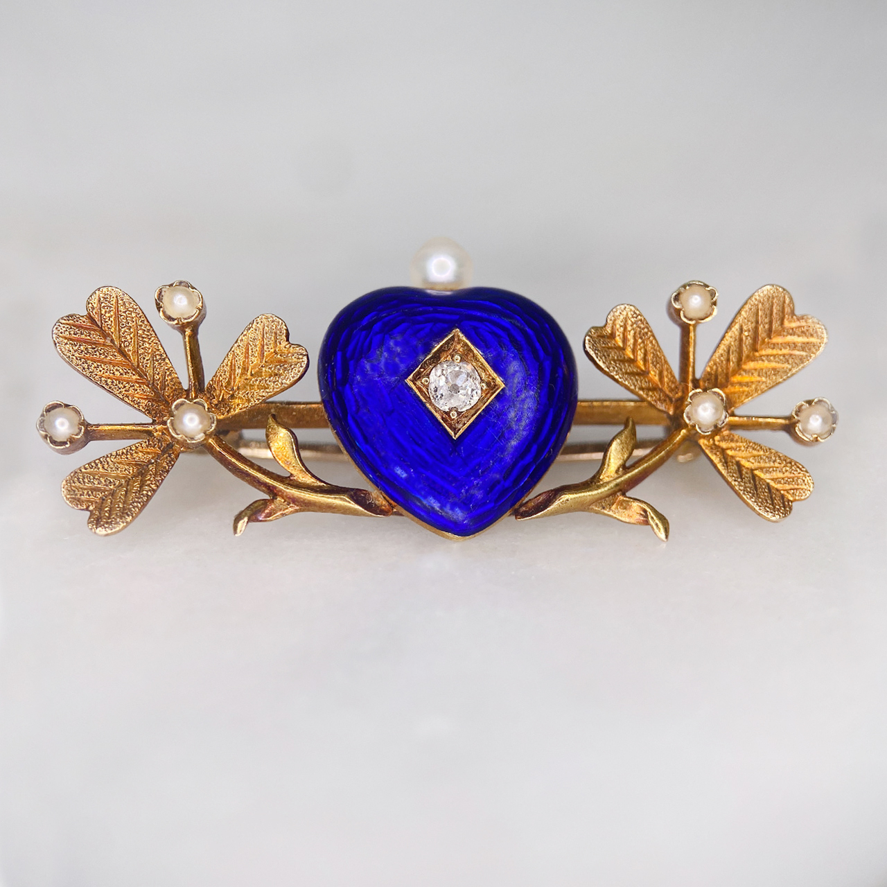 Much desired Victorian Blue Enamelled Heart and Clover Brooch, in 15  carat tested yellow gold.  Rich dark blue enamel heart with central old cut diamond and single pearl crown.  The heart is nestled between two pearl set clover leaves.  The clovers have chased leaf detail with seed pearl accents.  The whole brooch measures approximately 38mm x 16mm, all stones are claw set.  The rear of the brooch has a 15carat gold pin and C clasp with no safety catch.   The brooch would have possibly been a gift given from the heart to bring good luck to the recipient.  In immaculate condition, showing a small surface scratch to the bottom of the blue heart  in keeping with the age of the brooch.