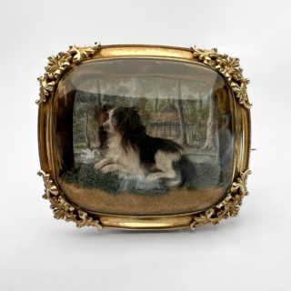 Rare Early Victorian Dog Model Brooch. Realistically modelled 3D King Charles spaniel mounted under slightly domed glass. The image is framed in 15ct yellow gold with beautiful scrolling foliage on the 4 corners. Behind the modelled dog there is a beautifully painted wood and lake scene with a swan and ornate building. The dog is resting on grass and sand in the foreground giving the image depth. The style of brooch was very popular in the Victorian period and represents the fascination of sentimentality as well as romanticised country life. The dog modelled in the brooch strongly resembles the beloved spaniels associated with Queen Victoria. The brooch measures at 53mm x 45mm. Please contact us if you would like to know more about this brooch or visit us at Saddingtons Jewellery, Mayfair.