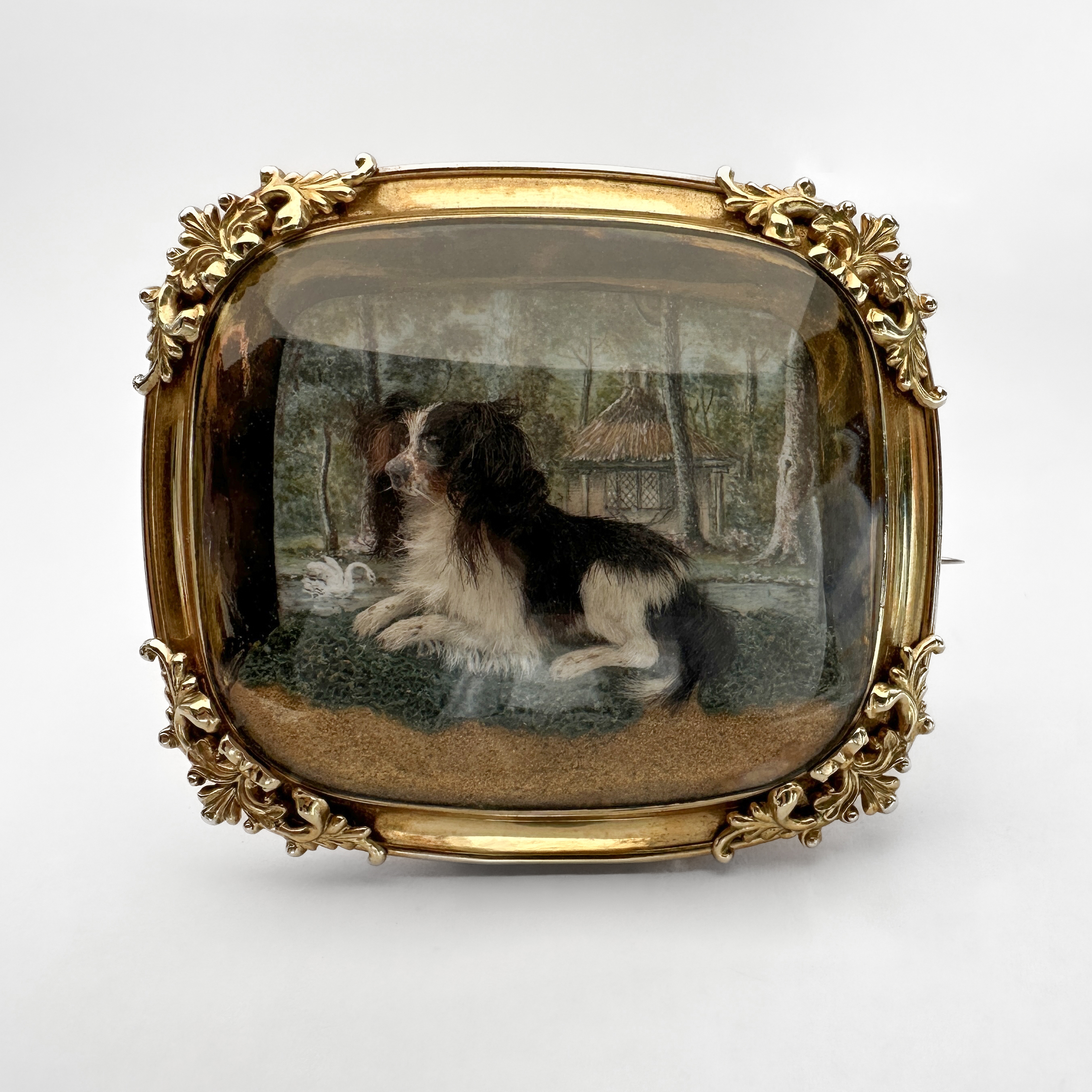 Rare Early Victorian Dog Model Brooch. Realistically modelled 3D King Charles spaniel mounted under slightly domed glass. The image is framed in 15ct yellow gold with beautiful scrolling foliage on the 4 corners. Behind the modelled dog there is a beautifully painted wood and lake scene with a swan and ornate building. The dog is resting on grass and sand in the foreground giving the image depth. The style of brooch was very popular in the Victorian period and represents the fascination of sentimentality as well as romanticised country life. The dog modelled in the brooch strongly resembles the beloved spaniels associated with Queen Victoria. The brooch measures at 53mm x 45mm. Please contact us if you would like to know more about this brooch or visit us at Saddingtons Jewellery, Mayfair.