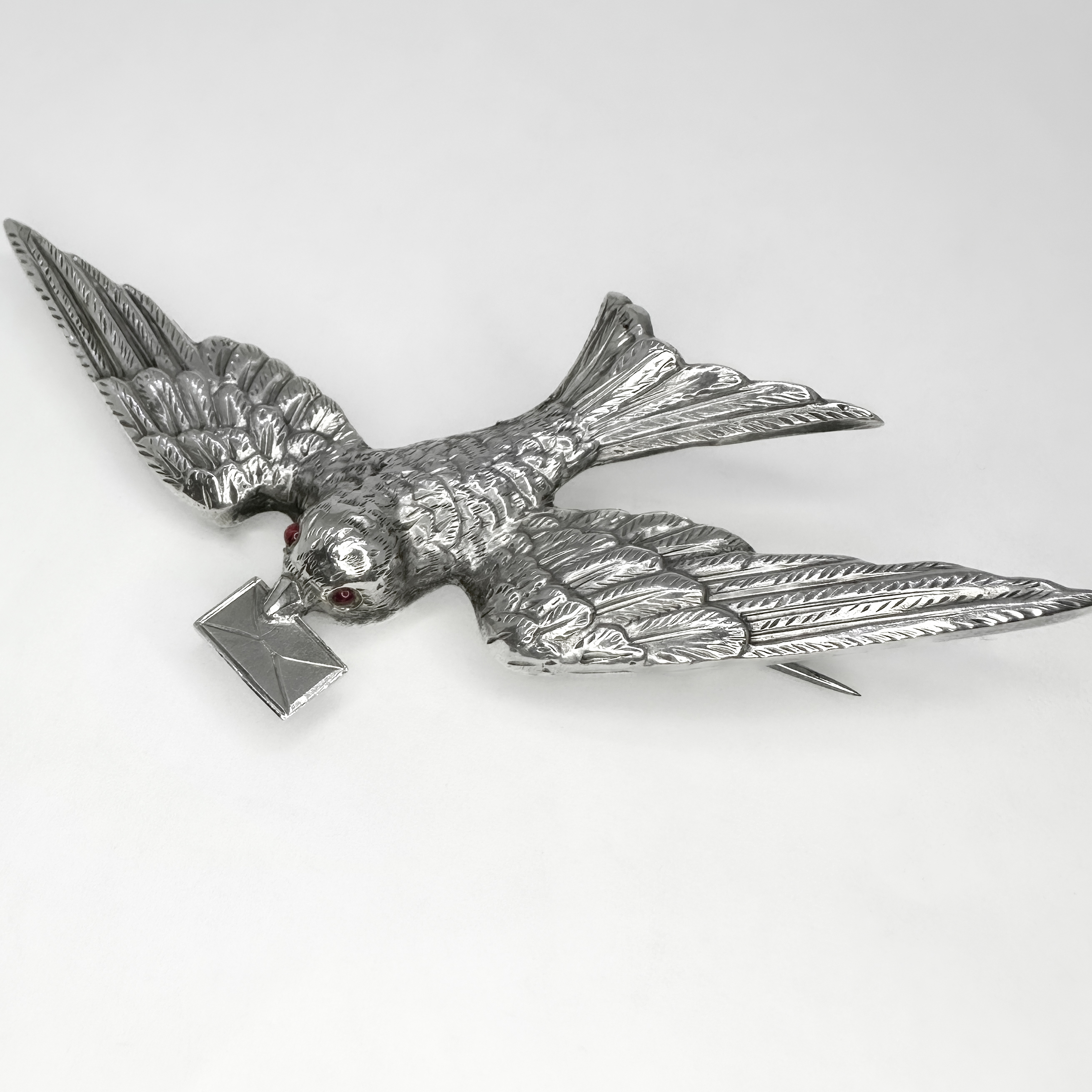Victorian Silver 'Love Bird' Brooch. The beautifully crafted silver chased swallow is captured in flight with a love letter in its beak, this is typical symbol of the victorian era representing love and hope. The silver carved bird has a hinged brooch pin securing on the back and features two small red pink rubies for its eyes. This romantic piece measures at 78mm x 45mm. Please contact us if you would like to know more about this brooch or visit us at Saddingtons Jewellery, Mayfair.