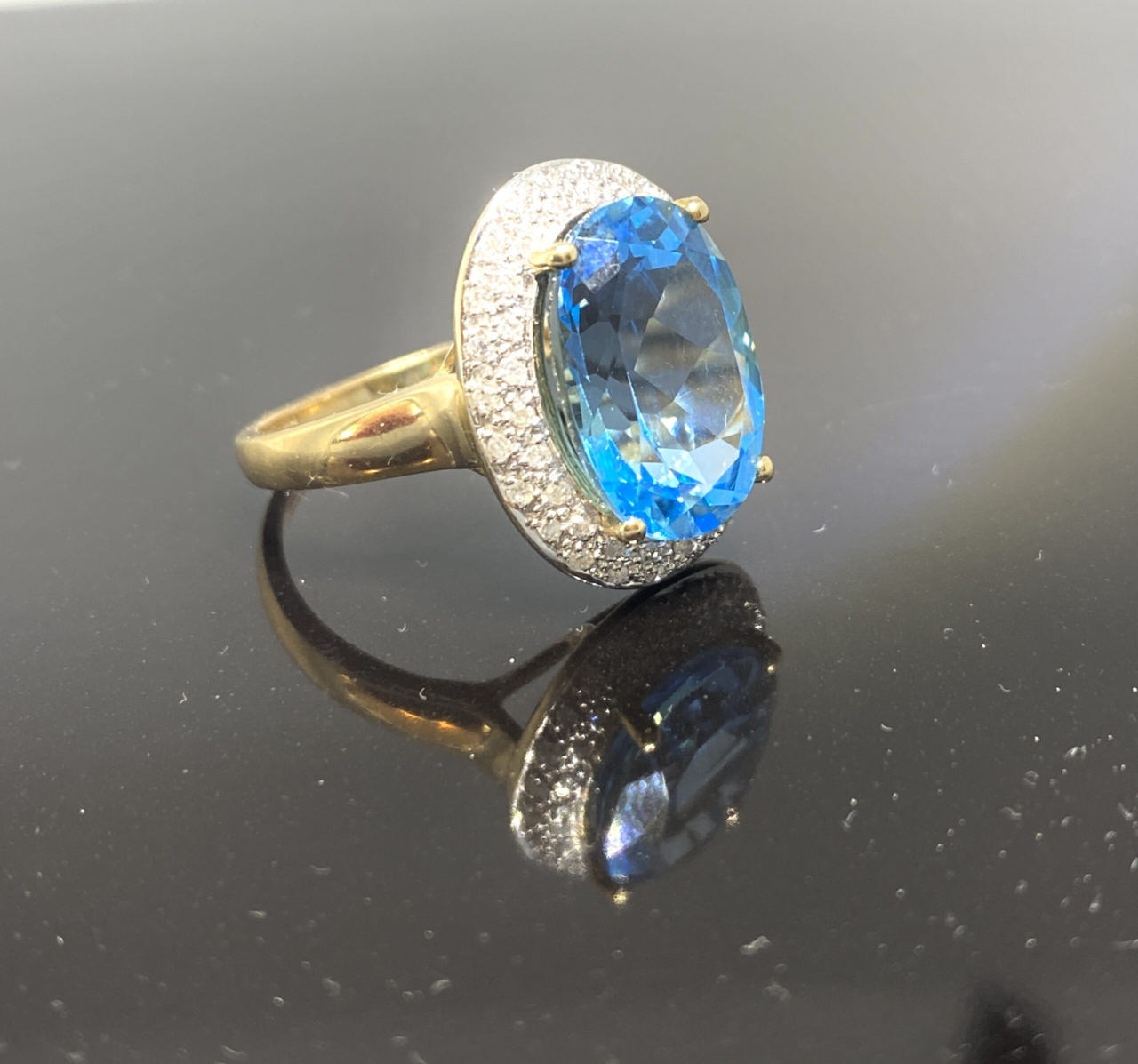 Blue Topaz Cocktail Ring 9ct, 6.5 carat blue topaz central stone surrounded by a halo of small sparkly diamonds.  The central blue topaz is beautifully faceted showing pale blue with glinting darker blue tones of summery swimming pool reminiscences!  A two row halo of small brilliant cut diamonds surround the blue topaz, an ideal elegant statement ring.  Comfortable to wear, a definite eye-catcher. The diamonds are pave set is in 9ct white gold on a 9ct yellow gold shank, fully hallmarked.  Message us or call to chat about the ring or to discuss your individual needs.