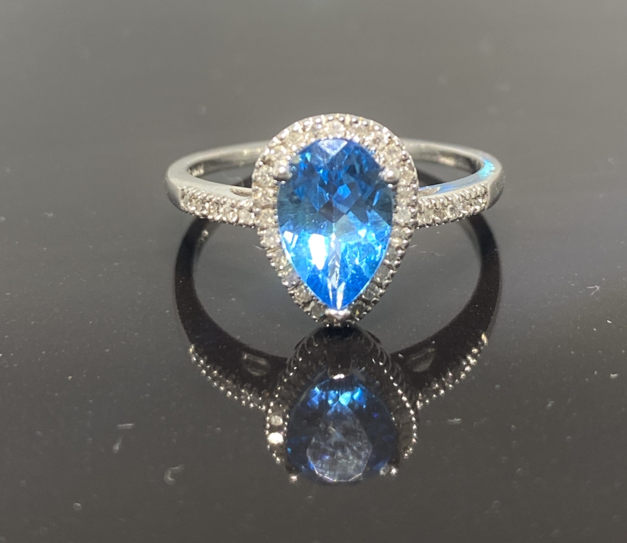 Blue Topaz Diamond Halo Ring, central pear shaped topaz surrounded with a halo of pave diamonds. The ring is in 10 ct white gold. The shoulders are set with pave diamonds also