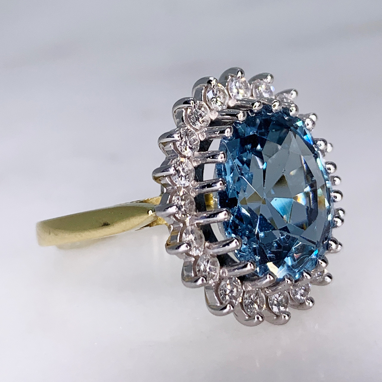 Aquamarine and Diamond Cluster Ring, 18ct Gold Hallmarked. The principle faceted Aquamarine and surrounding diamonds are claw set in a tiffany shank. The head of the ring measures approximately 18mm x 21mm.