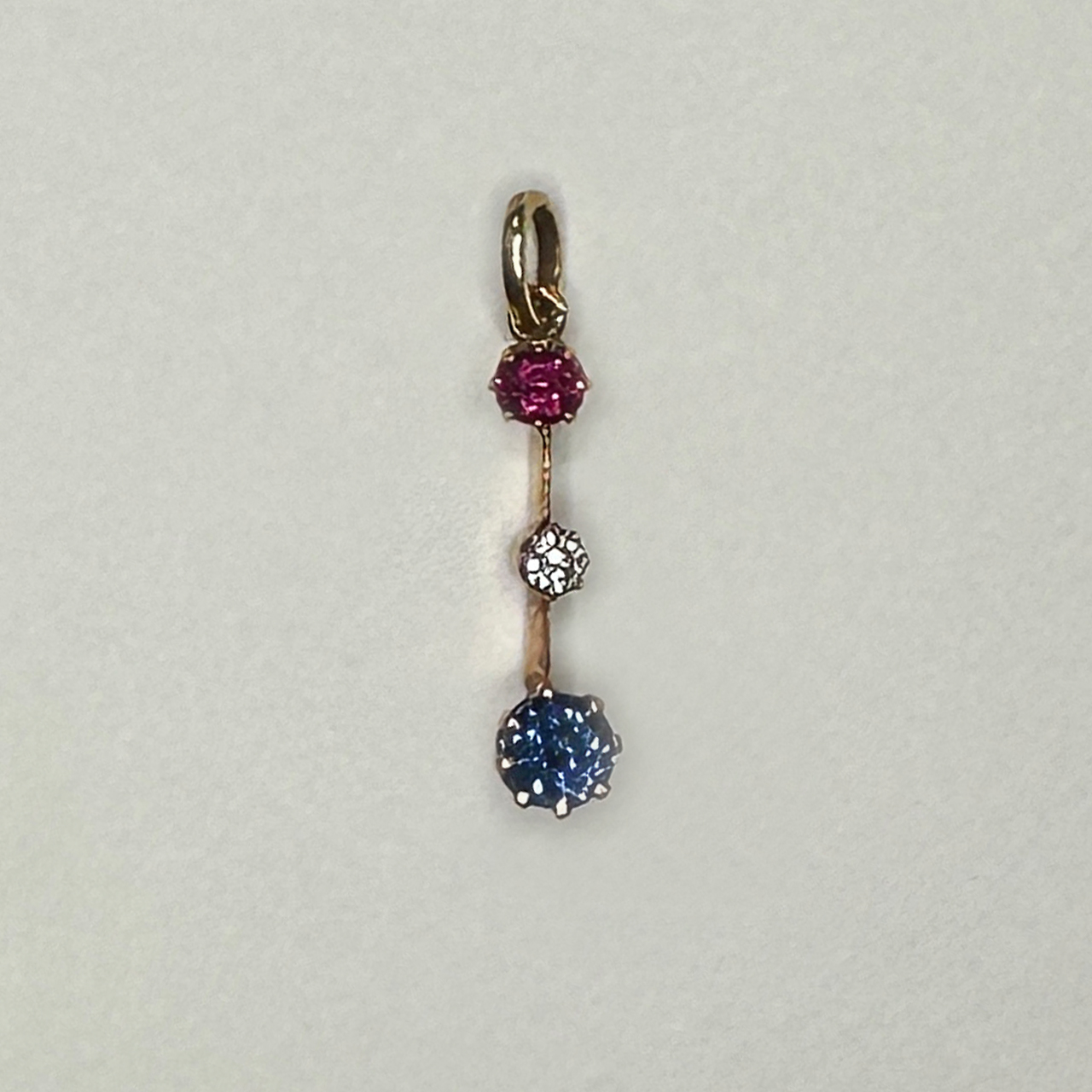 Exquisite fine Edwardian three stone gem pendant with colours red, white and blue. The pendant is set in a 15ct yellow gold. The ruby on top followed by a smaller brilliant cut antique diamond, the sapphire to the bottom of the pendant. The gems have claw settings. The drop including the bale is 2.5cm. 