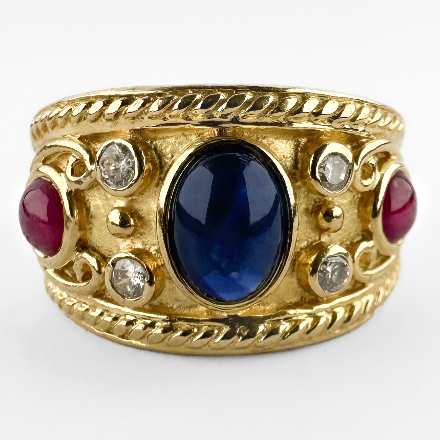 Wonderful Etruscan Cabochon Ruby & Sapphire Ring. A beautifully set rub over cabochon sapphire sits in the centre with 2 diamonds as accents and a stunning red round ruby on either side. The design is heavily influenced by classical Etruscan gold work, with intricate granulated detailing in the shank and scroll and rope sculptural forms that makes the ring look exceptional on the finger. The shank features an 18ct stamp alongside a GS makers mark and a continental assay mark. Likely of Swiss/Italian origin, circa 1970s–1980s, this is a beautifully made piece that reflects the high standard of continental gold-smithing of the period. Please contact us if you would like to know more about this incredible ring or visit us at Saddingtons Jewellery, Mayfair.