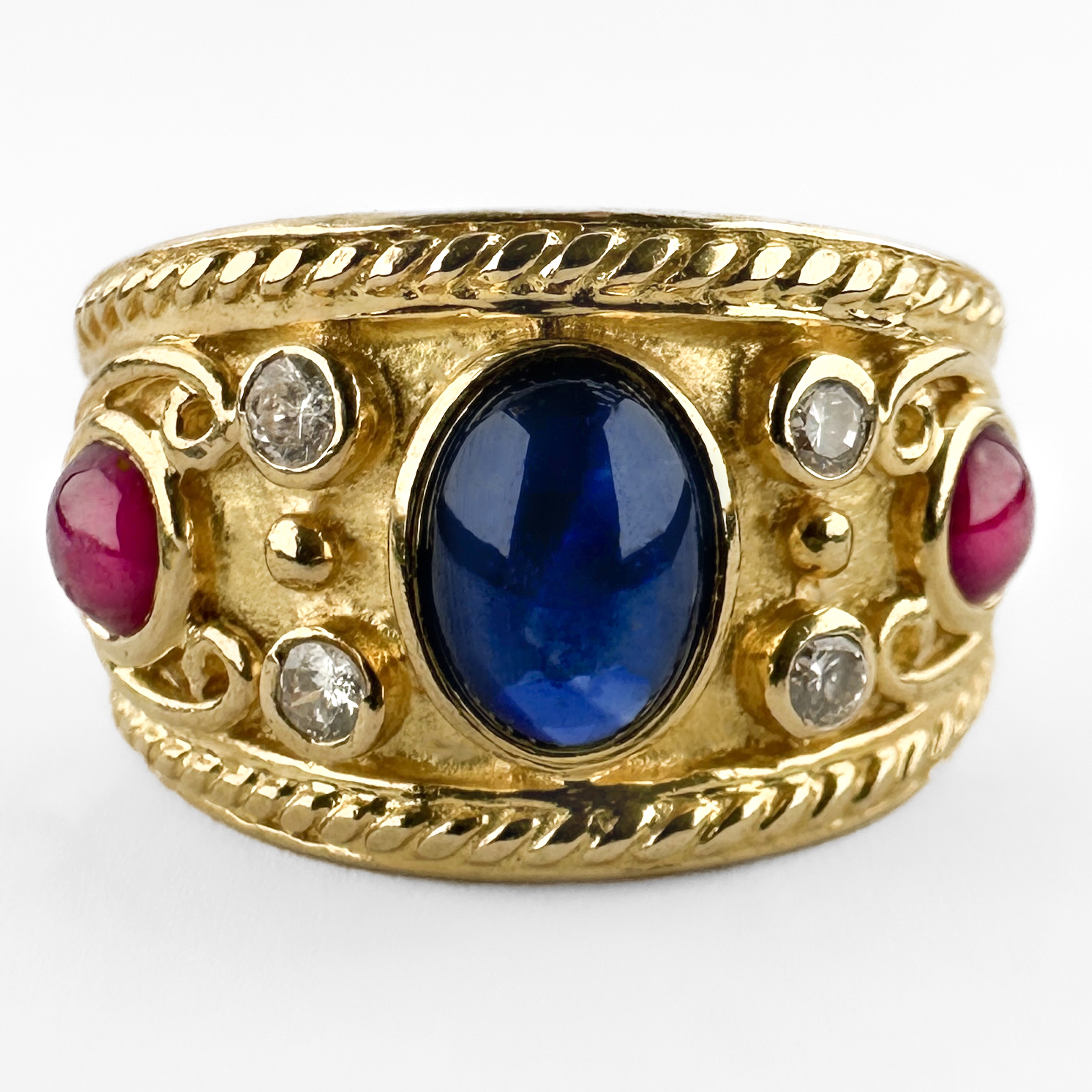 Wonderful Etruscan Cabochon Ruby & Sapphire Ring. A beautifully set rub over cabochon sapphire sits in the centre with 2 diamonds as accents and a stunning red round ruby on either side. The design is heavily influenced by classical Etruscan gold work, with intricate granulated detailing in the shank and scroll and rope sculptural forms that makes the ring look exceptional on the finger. The shank features an 18ct stamp alongside a GS makers mark and a continental assay mark. Likely of Swiss/Italian origin, circa 1970s–1980s, this is a beautifully made piece that reflects the high standard of continental gold-smithing of the period. Please contact us if you would like to know more about this incredible ring or visit us at Saddingtons Jewellery, Mayfair.