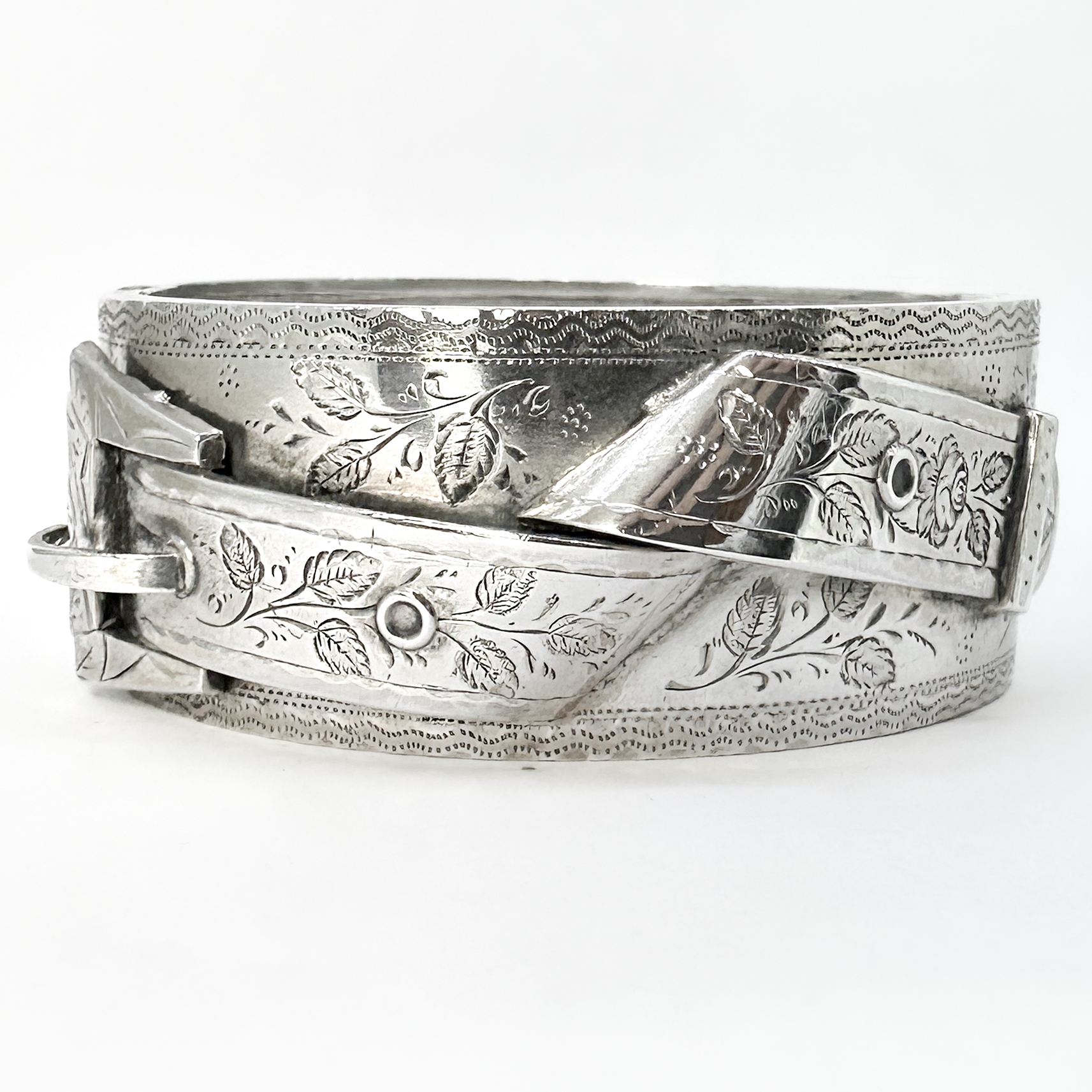 Wonderful Victorian Silver Belt Cuff Bracelet. A beautifully engraved Victorian sterling silver cuff bracelet dated to 1886, hallmarked with a Birmingham assay mark. This striking wide-form bracelet is decorated throughout with elegant hand-engraved floral and foliate motifs showcasing the refined artistry of late 19th century silver work. The bracelet features a crossover buckle-inspired design to the front, combined with a patented 'west patent' sprung interior construction which allows the piece to sit comfortably and securely on the wrist. The rich engraving and softly aged silver patina give the bracelet exceptional depth and character. Both highly wearable and distinctly decorative, this is a wonderful example of substantial Victorian silver jewellery — elegant enough for everyday wear yet bold enough to make a statement. A rare and beautifully preserved antique silver cuff with timeless appeal. Please contact us if you would like to know more about this amazing bracelet or visit us at Saddingtons Jewellery, Mayfair.