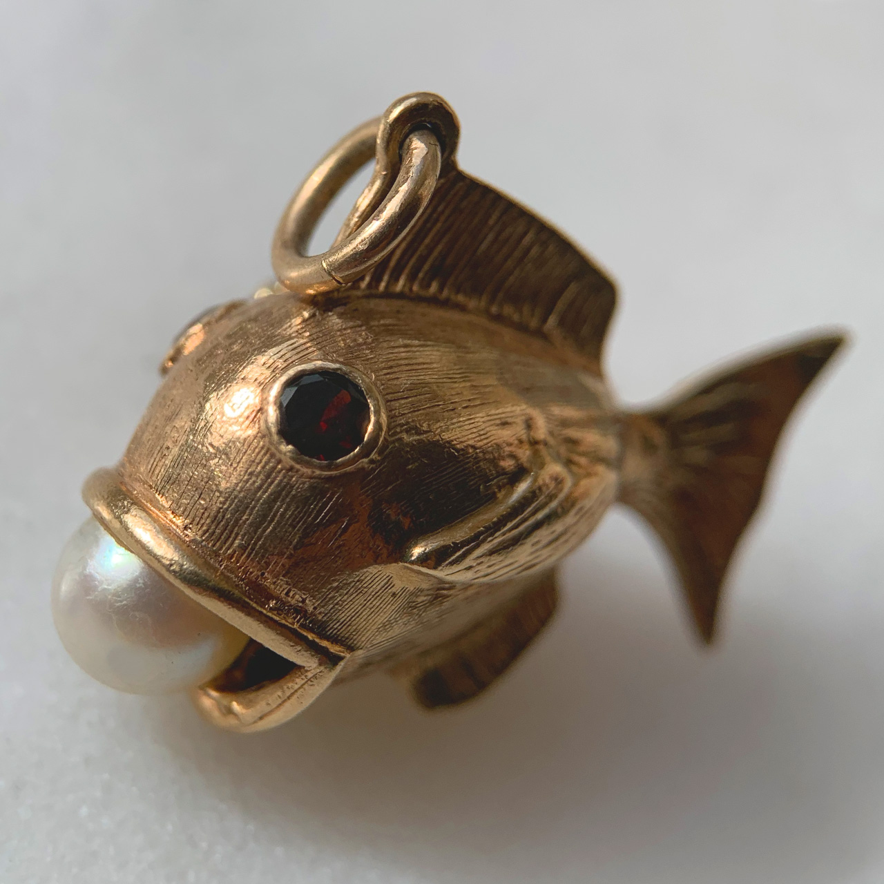 Characterful Vintage Garnet and Pearl in Mouth Fish Charm in 9ct Rose Gold, Hallmarked London, 1965. This wonderful charm has two deep garnet eyes, bezel set, measuring approximately 3 mm in diameter. The fish holds a fresh water pearl 6 mm in diameter in an open mouth. The fish is designed with intricate fin and scale chased detail. The total fish measures 31 mm x 22 mm (excluding the 7 mm loop). The charm is in excellent condition and a perfect quirky addition to any charm bracelet or necklace.