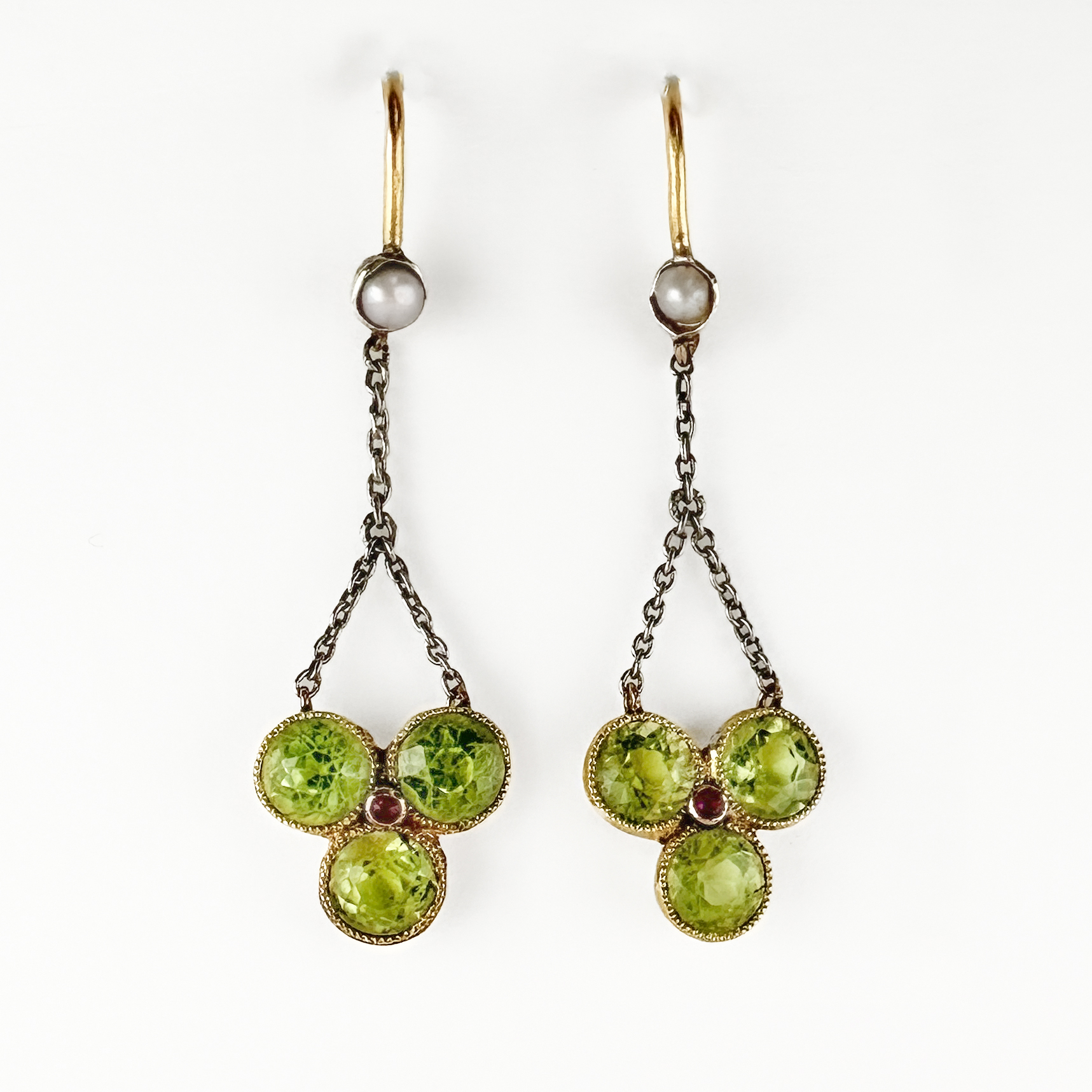 Sweet Antique Period Trefoil Drop Earrings. These beautiful late victorian have three gorgeous green peridots in a trefoil 'clover' formation with a central small ruby this shape is suspended from a 15ct chain. At the top of the chain there is a rub over set natural pearl. The earrings have shepherd's crook ear fittings also in 15ct yellow gold. The drop from the pearl to end measures at 2.5inches. Please contact us if you would like to know more about these rare and pretty earrings or visit us at Saddingtons Jewellery, Mayfair.