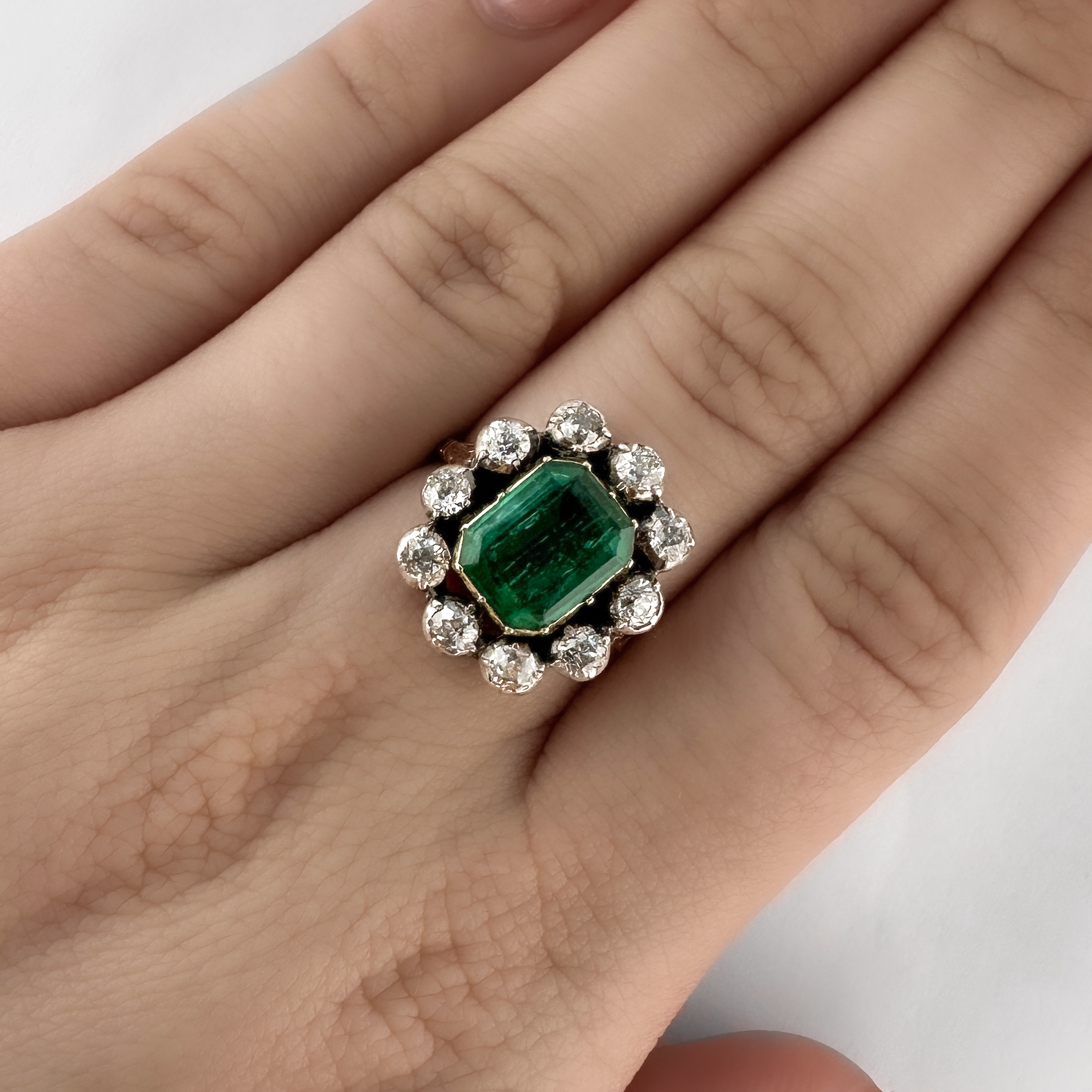 Stunning Victorian Emerald & Diamond Cluster Ring. This ring features a central rectangular natural emerald surrounded by 10 old cut diamonds. The ring mount is carved and the stones are all set closed back in 18ct yellow gold which is very typical of the era. The head of the ring measures at 1.8cm x 1.6cm. Please contact us if you would like to know more about this stunning ring or visit us at Saddingtons Jewellery, Mayfair.