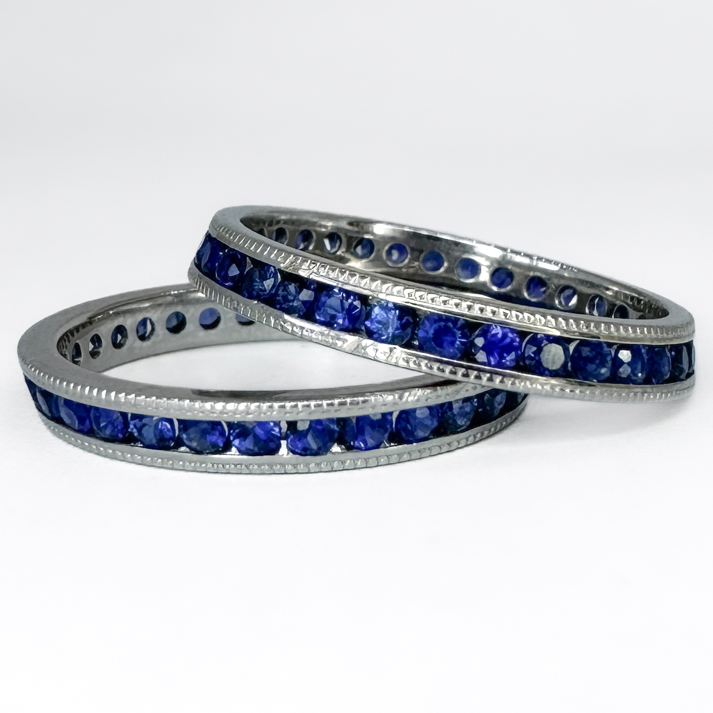 A Pair of Stacking Sapphire Eternity Rings. Both full eternity ring includes round cut royal blue sapphires that are channel set in 18ct white gold. The pair have a slight slope to their edges that make them perfect for stacking and other rings to sit between. They also feature millegrain detailed edging and are marked 1950s. The rings measure at  6mm in width when stacked on top of each other. As the rings are full sapphire ring bands, they cannot be resized. Please contact us if you would like to know more about these rings or visit us at Saddingtons Jewellery, Mayfair.
